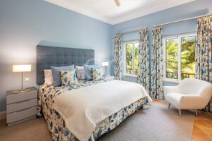 Blue bedroom with a tufted headboard bed, floral bedding, two nightstands, and a white armchair beside large windows with patterned curtains.