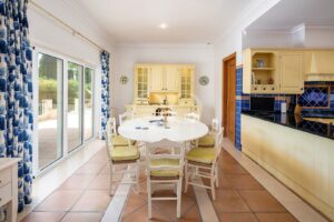 Sunlit dining area with a round white table and cream chairs, blue tile kitchen to the right, and sliding glass doors with blue patterned curtains opening to an outdoor patio