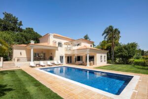 Cream-colored villa with terracotta roof, outdoor pool, and sun loungers on a tiled patio under a clear blue sky.