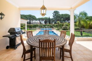 Round wooden dining table with six chairs on a tiled patio, a gas grill on the left, overlooking a rectangular pool and lush garden beyond.