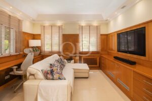 Living room with built-in wooden cabinetry, a large flat-screen TV, a white sofa with floral cushions, and windows with wooden blinds.