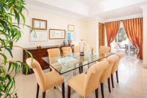 Bright dining room with glass table, beige checkered chairs, framed certificates on the wall, and orange curtains at the patio door.