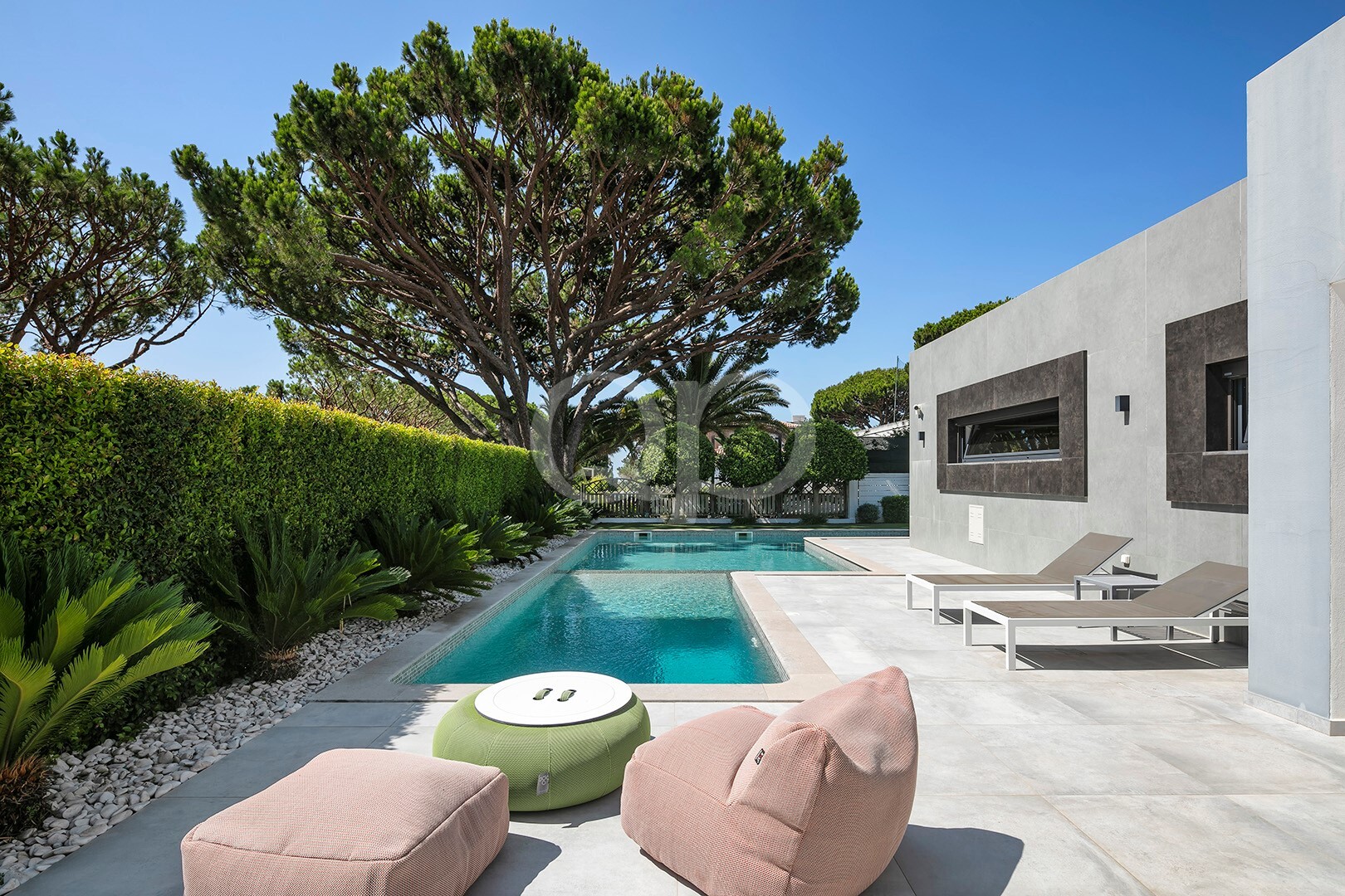 Un jardin moderne avec une piscine rectangulaire, des chaises longues couleur beige et des haies soigneusement taillées, le tout sous un ciel bleu.