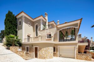 Front view of a cream Mediterranean villa with stone base, tiled roof, and multiple balconies under a bright blue sky