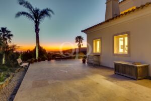 Vista do pôr-do-sol a partir de um pátio com azulejos, junto a uma casa bege com janelas iluminadas e palmeiras. Um banco, vasos com plantas e mobiliário de exterior encontram-se ao longo da borda.