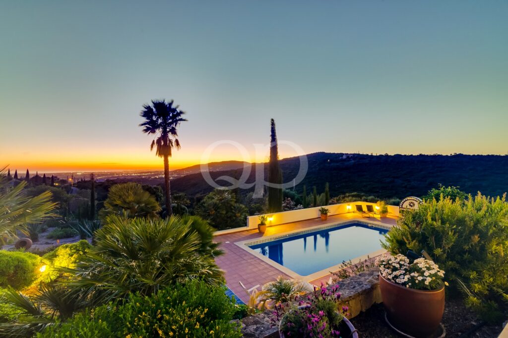 Piscina no terraço ao anoitecer, com iluminação acolhedora, plantas em vasos e um pôr-do-sol ao longe, sobre a encosta.