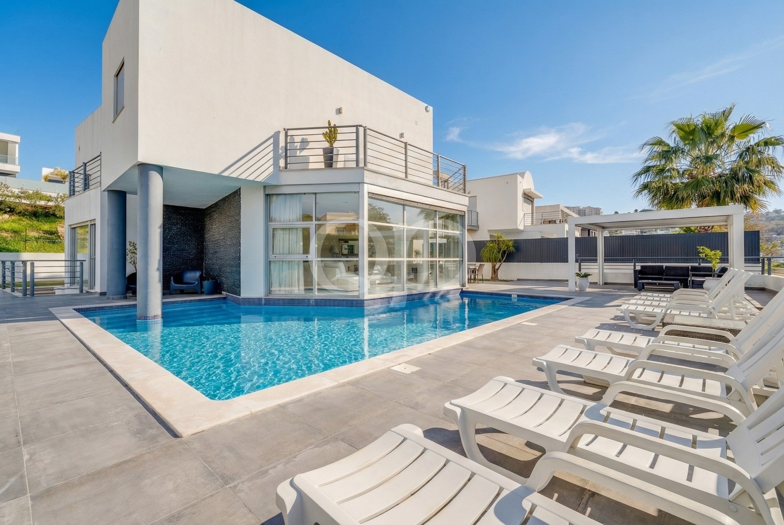 Moradia moderna branca com uma piscina retangular azul, uma sala de estar com paredes de vidro e uma fila de espreguiçadeiras brancas sob um céu ensolarado.