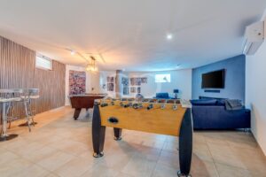 Sala de jogos na cave com matraquilhos e mesas de bilhar, bancos de bar à esquerda e um sofá azul ao longo da parede da direita.