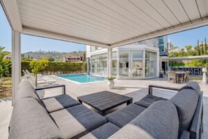 Área de estar exterior coberta com vista para uma piscina retangular e para uma casa moderna com paredes de vidro.