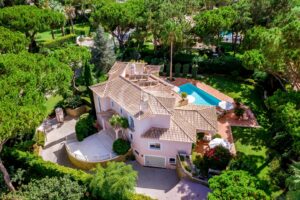 Vista aérea de uma moradia em estuque cor-de-rosa com telhado de terracota, rodeada por árvores frondosas e uma piscina retangular azul num jardim ajardinado.]','Imagem aérea de uma moradia de estilo mediterrânico com telhas de terracota, paredes cor-de-rosa e uma piscina adjacente a um pátio de terracota.','Luxuosa moradia na encosta, emoldurada por árvores altas, com telhado de telhas vermelhas, exterior rosa e uma ampla área de piscina.','Vista aérea de uma propriedade ajardinada com uma casa central, telhado de terracota e piscina azul brilhante no meio de uma vegetação exuberante.','Moradia vista de cima: exterior rosa, telhado de telhas de barro, jardim bem cuidado e um pátio com assentos ao ar livre junto à piscina.