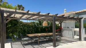 Outdoor dining area under a wooden pergola with a long table and metal chairs, surrounded by green bushes and a white building. It’s sunny with clear blue sky.