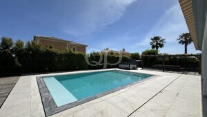 Rectangular pool with turquoise water bordered by light stone tiles, patio seating area in the background under a clear blue sky.