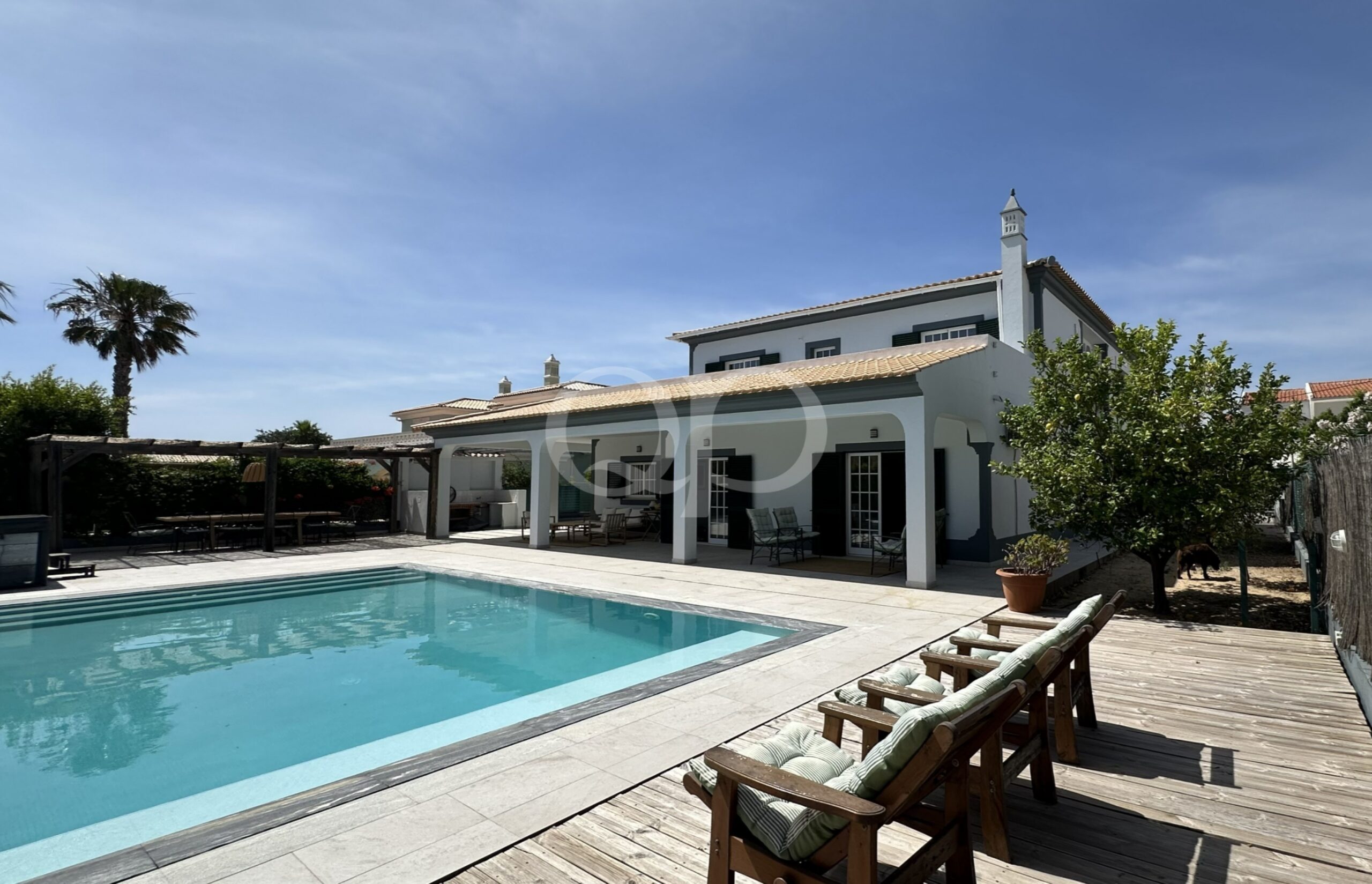 Espace piscine dans le jardin, avec une piscine rectangulaire de couleur turquoise, des chaises longues en bois et une maison blanche voûtée, le tout sous un ciel bleu ensoleillé.