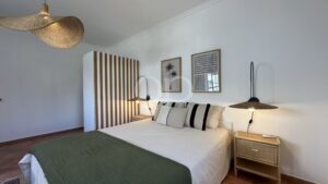 Bedroom with white bedding and a green quilt, wicker pendant lamps, striped room divider, and palm-framed wall art on white walls.
