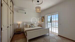 Bright bedroom with white wooden bed, matching nightstands, and built-in wardrobes on the left; large window fills the room with light.