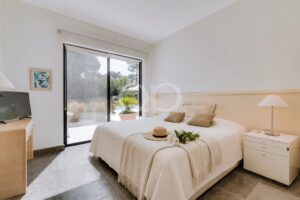 Quarto ao estilo de hotel com uma cama queen size, roupa de cama branca e almofadas bege, duas mesas de cabeceira e uma porta de vidro deslizante que dá acesso a um pátio com jardim.
