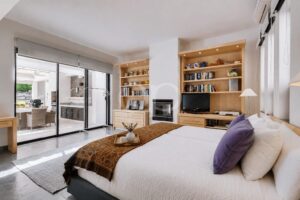 Quarto luminoso com roupa de cama branca, almofadas roxas e prateleiras de madeira embutidas de ambos os lados da lareira e da televisão; as portas de vidro de correr dão acesso a um pátio.
