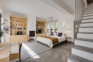 Estúdio luminoso e arejado com uma cama king size, cabeceira bege e almofadas roxas, sob um teto branco e grandes janelas