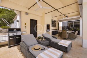 Pátio exterior com paredes bege, duas espreguiçadeiras de vime, um grelhador embutido e uma mesa de jantar sob um telhado coberto com ventiladores.