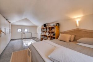 Quarto no sótão com uma cama grande e cabeceira de madeira, roupa de cama bege e uma estante alta ao longo da parede.