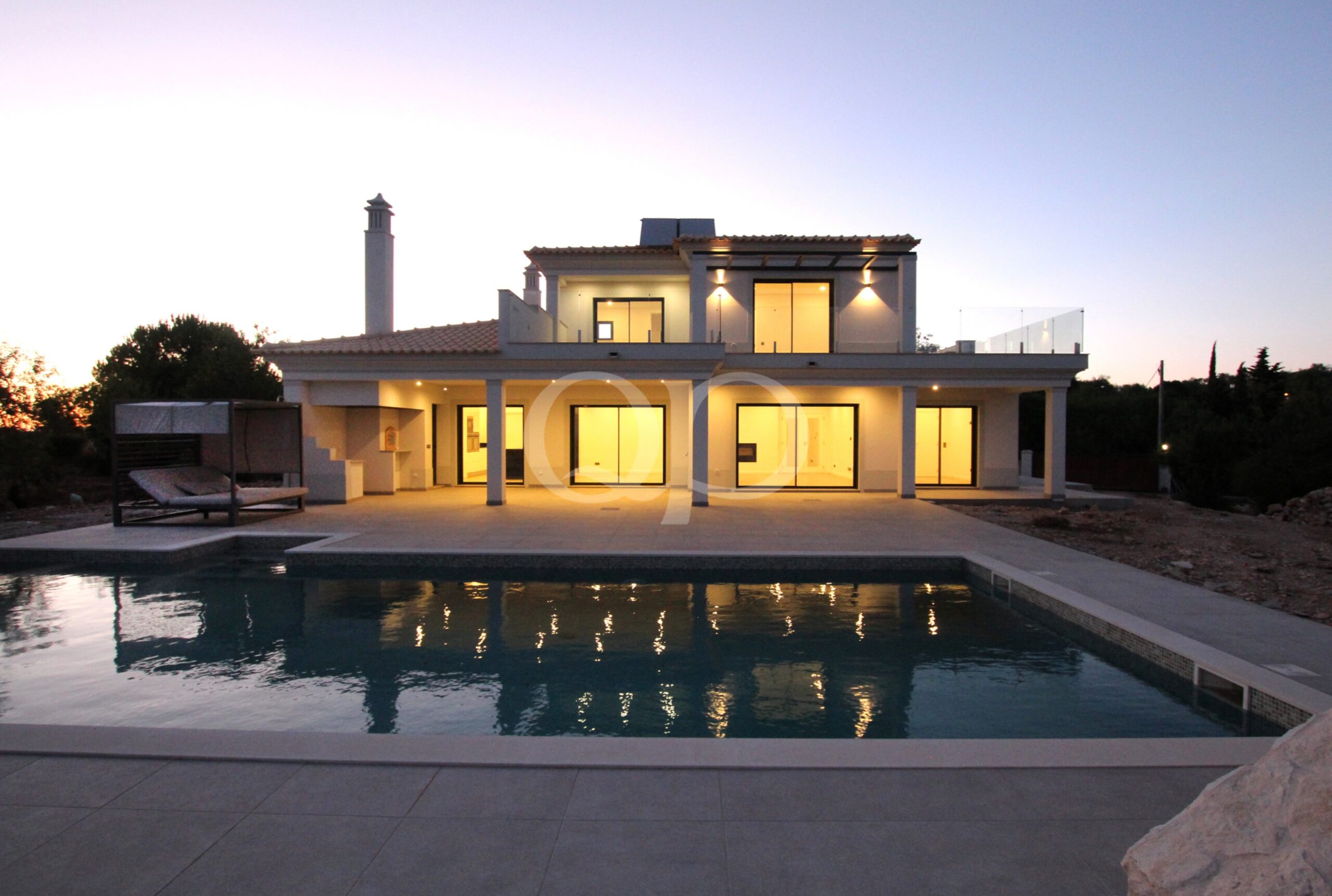 Casa branca moderna de dois andares, com grandes portas de vidro, luzes interiores acolhedoras e uma piscina iluminada em primeiro plano ao anoitecer.