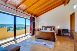 Quarto deslumbrante e espaçoso, com piso e teto em madeira, dotado de amplas portas de vidro que dão acesso a uma varanda privada com vistas panorâmicas sobre a paisagem rural