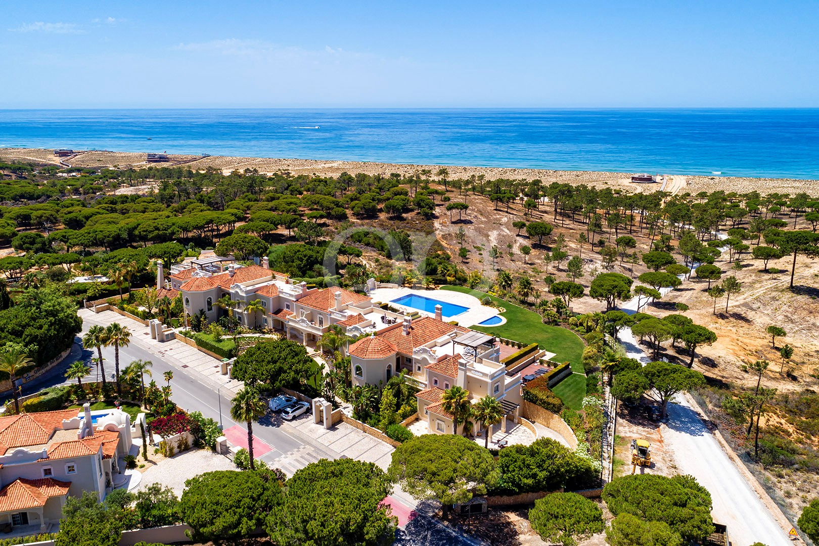 A Mediterranean-style villa complex with red-tiled roofs, a central blue pool, palm trees, and a coastal sea backdrop.
