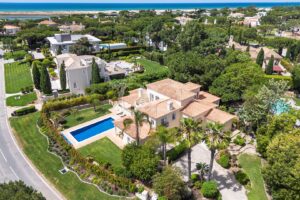 Aerial view of a luxury coastal neighborhood with beige villas, terracotta roofs, palm trees, and a blue rectangular pool surrounded by greenery near the shore.
