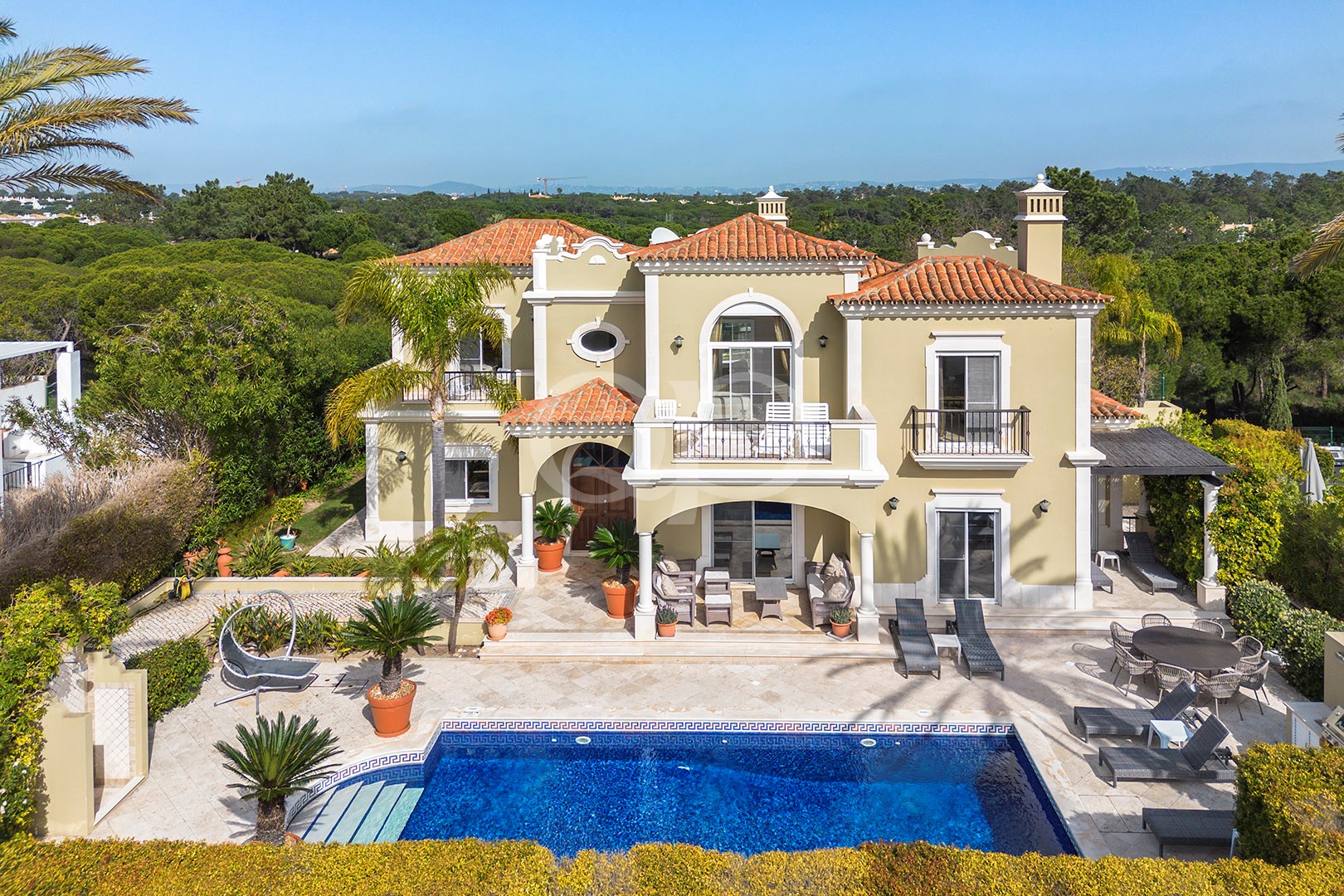 Luxuriöse beigefarbene Villa im mediterranen Stil mit rotem Ziegeldach, Balkon und einem blauen, rechteckigen Swimmingpool im Vordergrund.