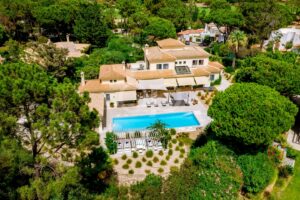 Vista aérea de uma moradia de estilo mediterrânico com telhados de terracota, paredes bege e uma longa piscina azul rodeada por espreguiçadeiras e um pátio à sombra.