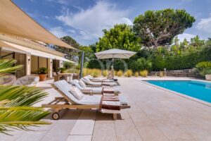 Pátio de luxo junto à piscina, com espreguiçadeiras almofadadas, toalhas e um grande guarda-sol junto a uma piscina retangular azul.