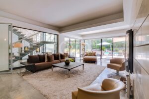 Sala de estar em plano aberto com um sofá seccional castanho, uma mesa de centro preta e um tapete felpudo; as paredes de vidro oferecem vista para a zona da piscina no exterior.
