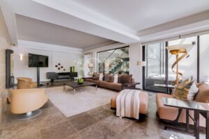 Sala de estar em plano aberto com um sofá castanho comprido, cadeiras bege e uma parede de portas de vidro que dá para uma escadaria no exterior. Continue a ler: a luz encontra o design contemporâneo.