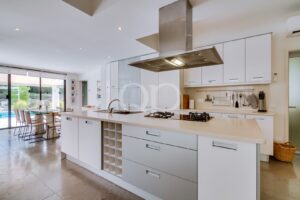 Cozinha em plano aberto com uma grande ilha branca, exaustor em aço inoxidável e eletrodomésticos embutidos, que dá acesso a uma zona de refeições com uma mesa de vidro e vista para a piscina exterior