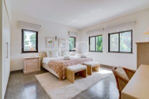 Quarto luminoso com uma cama queen size, colcha com motivos florais, dois bancos de vime aos pés da cama e mesas de cabeceira de madeira clara junto a grandes janelas com persianas brancas.