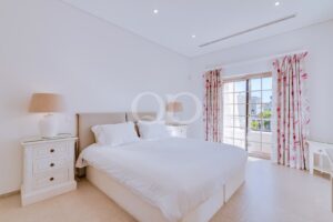 Bright hotel bedroom with a large white bed, beige headboard, two white nightstands, and lamps, plus pink floral curtains framing a glass door to the balcony.