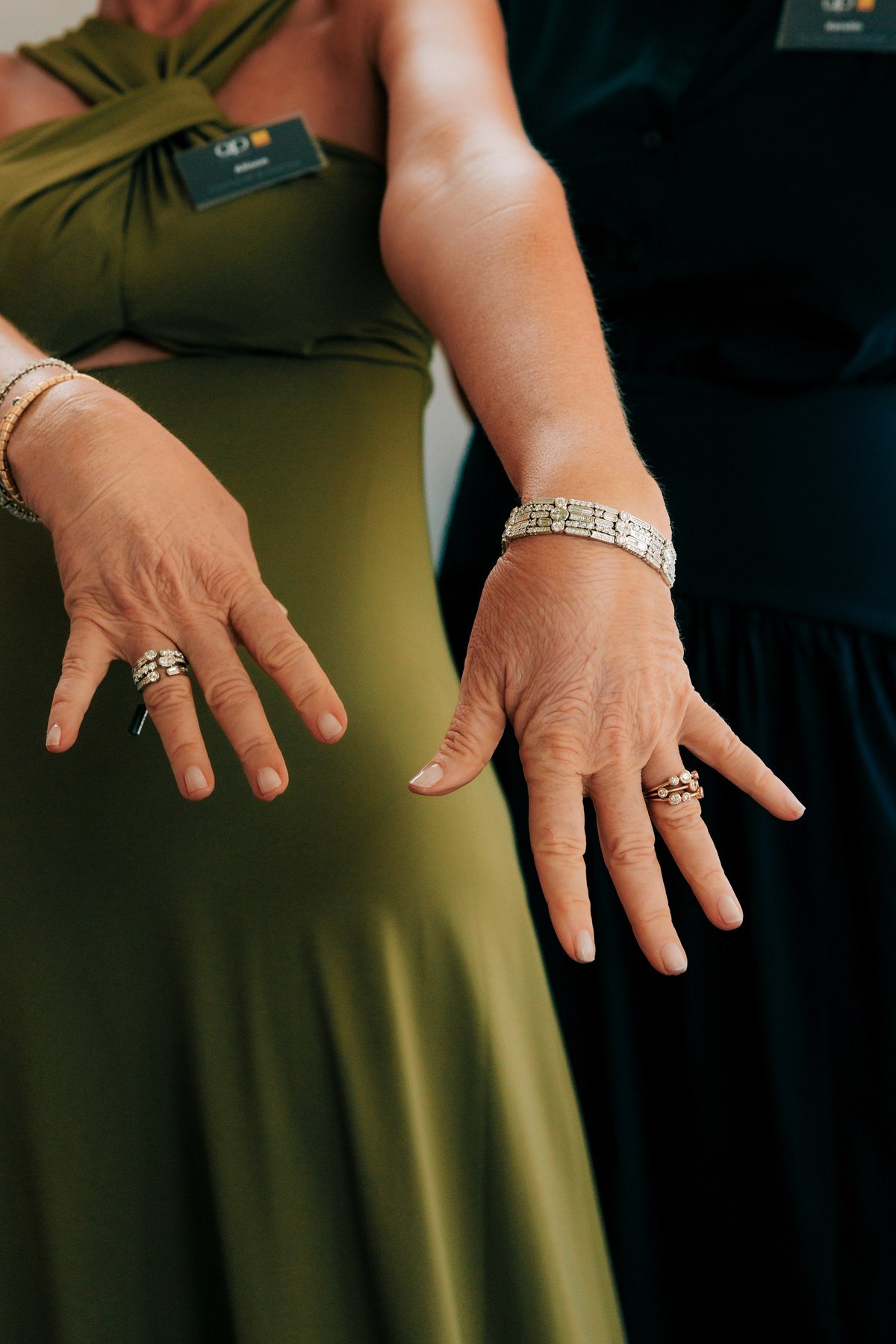 Die Hände zweier Frauen zeigen ihren Schmuck: Die eine trägt einen Diamantring und ein Armband an einem Arm, der in Grün gekleidet ist, die andere mehrere Ringe und ein Armband, die sich von ihrem dunklen Outfit abheben.